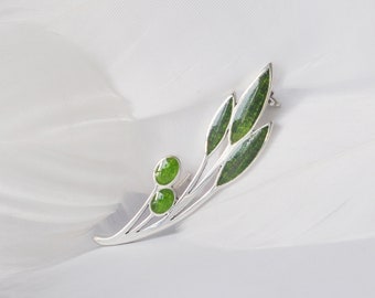 May include: A silver brooch in the form of a stylized leaf branch. The brooch has three elongated green leaves and two round green accents. The brooch is set against a white background.