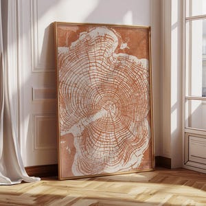 May include: A framed art print with a tree cross-section design. The artwork shows concentric rings in white against a terracotta background. The print is in a gold frame and leans against a white wall. The print is in a gold frame.