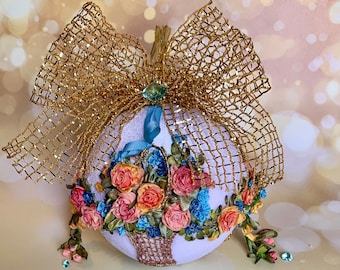 May include: A white Christmas bauble with a gold mesh bow and a floral design made of ribbon. The bauble features a basket filled with pink, peach, and blue ribbon flowers.