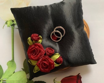 May include: Black satin ring pillow with red satin roses and green leaves. Two silver wedding bands are placed on the pillow.