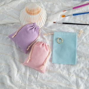 Puede incluir: Tres pequeñas bolsas de cordón de tela. Las bolsas son de color lavanda, rosa y azul claro. Un anillo de oro está dentro de la bolsa azul claro.