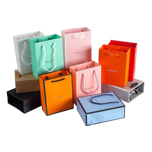 May include: Assortment of colorful paper gift bags with handles. The bags are decorated with the word "Viniknde" in a gold script font. The bags are arranged in a variety of colors, including pink, blue, green, orange, and red.