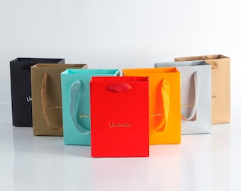 May include: A set of six colorful paper gift bags with handles. The bags are black, brown, light blue, red, orange, and gray. Each bag has the word "remarkable" printed on the front in gold lettering.