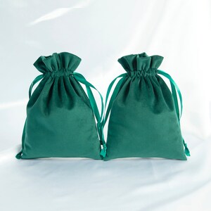 Pode incluir: Duas bolsas de cord&atilde;o de veludo verde com la&ccedil;os de fita de cetim combinando. As bolsas est&atilde;o vazias e colocadas sobre uma superf&iacute;cie branca.