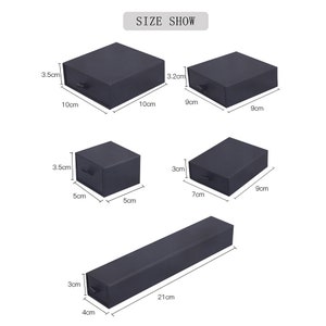 Op de afbeelding: Vier zwarte dozen met laden in verschillende maten. De dozen meten 10 cm x 10 cm, 9 cm x 9 cm, 5 cm x 5 cm en 7 cm x 9 cm. Een lange zwarte doos meet 21 cm x 4 cm.