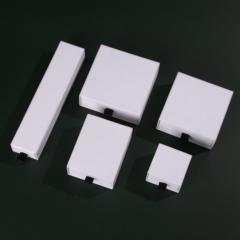 May include: Five white cardboard boxes with black pull tabs. The boxes are of different sizes and are arranged in a staggered pattern on a green surface.