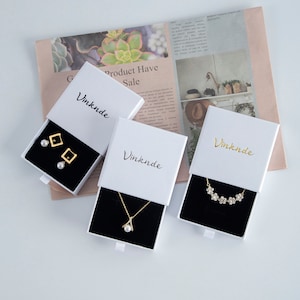 Op de afbeelding: Drie witte geschenkdozen met een zwart fluwelen interieur, elk met een ander sieraad. De dozen zijn gelabeld met "Vinknde" en hebben een schuiflade. De sieraden omvatten een paar parel oorbellen met gouden vierkante accenten, een gouden ketting met een parel hanger en een gouden ketting met een bloemenpatroon.