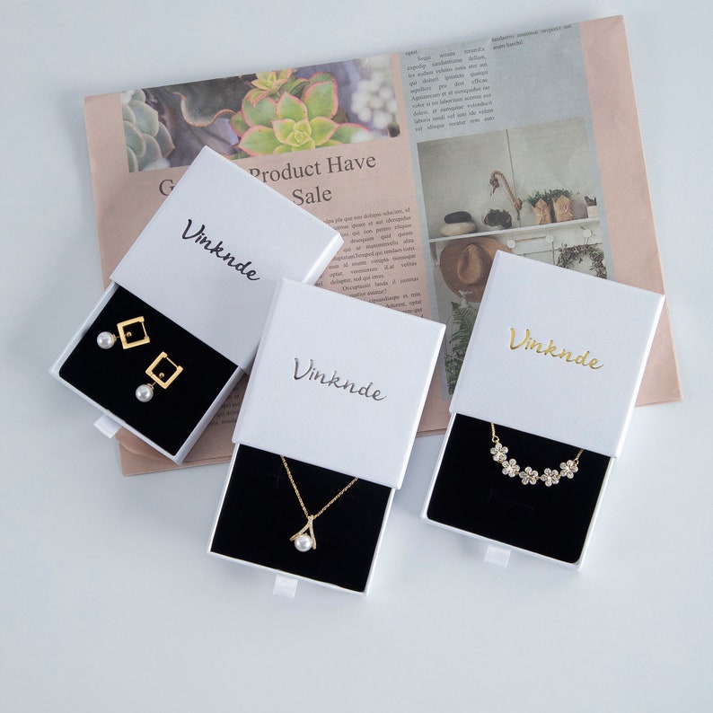 Op de afbeelding: Drie witte geschenkdozen met een zwart fluwelen interieur, elk met een ander sieraad. De dozen zijn gelabeld met "Vinknde" en hebben een schuiflade. De sieraden omvatten een paar parel oorbellen met gouden vierkante accenten, een gouden ketting met een parel hanger en een gouden ketting met een bloemenpatroon.
