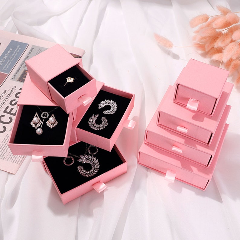 May include: Five pink stackable jewelry boxes with drawers. Each box has a black velvet interior and is decorated with a satin ribbon. The boxes are filled with various jewelry, including earrings, necklaces, and rings.