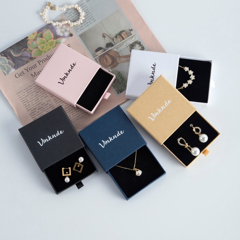 Op de afbeelding: Vijf kleine geschenkdozen met laden, elk in een andere kleur: roze, wit, bruin, zwart en blauw. Op elke doos staat het woord "Vinknde" in witte letters gedrukt. De dozen bevatten verschillende sieraden, zoals kettingen, oorbellen en armbanden.