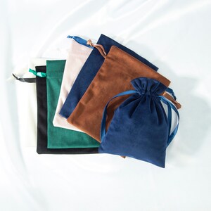 Pode incluir: Cinco sacos de veludo com cord&atilde;o de ajuste em diferentes cores, incluindo branco, verde, rosa, marrom e azul. Os sacos est&atilde;o empilhados, com o saco branco na parte inferior e o saco azul na parte superior.