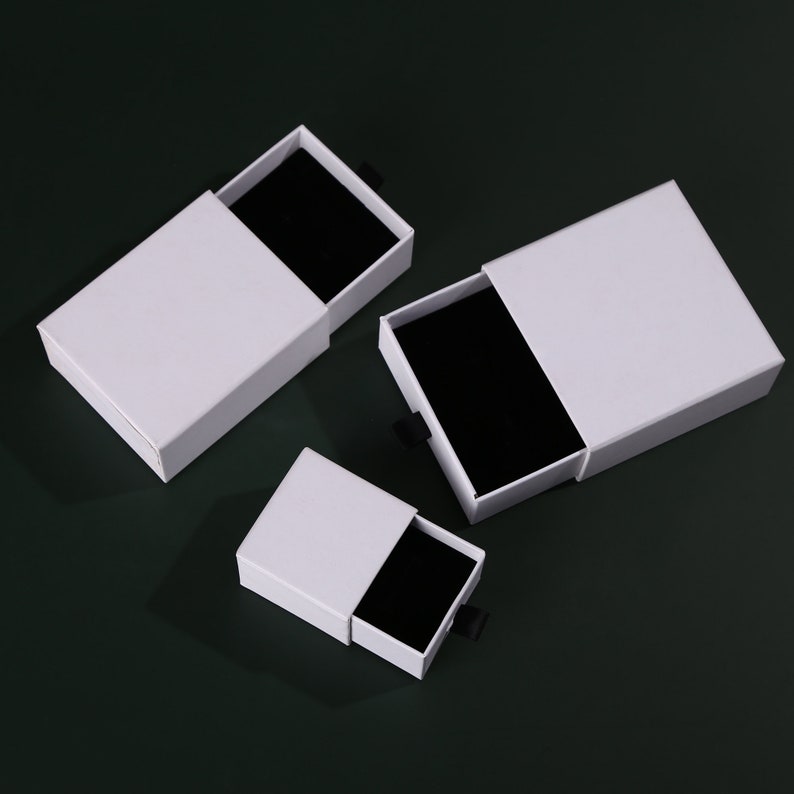 May include: Three white gift boxes with black velvet interiors and black pull tabs. The boxes are of different sizes and are arranged on a dark green surface.