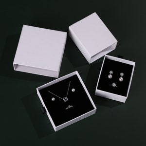 May include: Three white gift boxes with black velvet interiors. The boxes contain jewelry, including a necklace with a blue stone pendant, a pair of pearl earrings, and a ring.