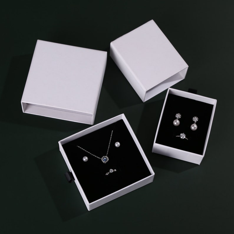 May include: Three white gift boxes with black velvet interiors. The boxes contain jewelry, including a necklace with a blue stone pendant, a pair of pearl earrings, and a ring.