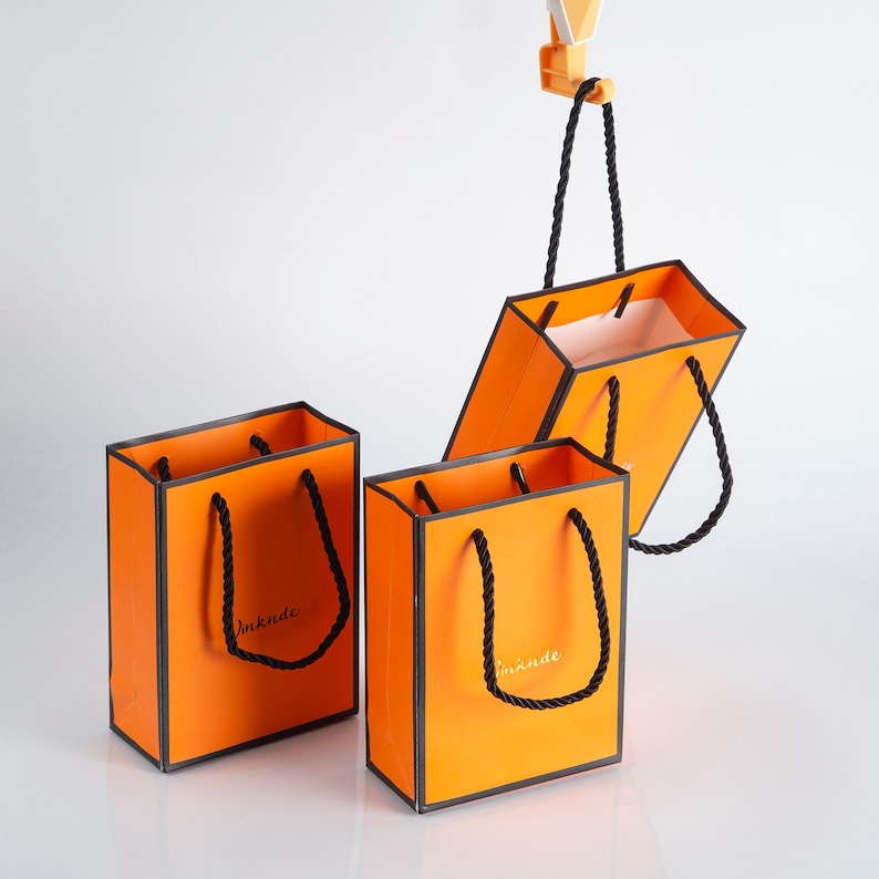 May include: Three orange gift bags with black handles and black trim. The bags have the text "imknde" printed on them.