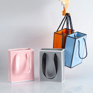 May include: Three gift bags in different colors: pink, gray, and blue. The bags have white trim and black handles. The blue bag has a black rope handle.
