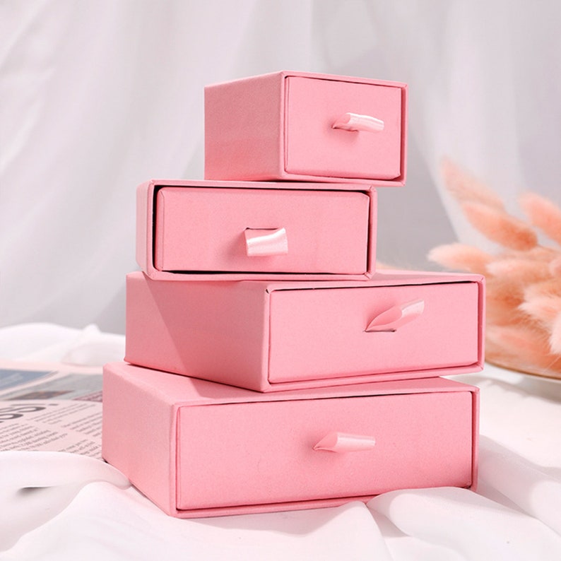 May include: A stack of four pink drawer boxes with satin ribbon pulls. The boxes are nested inside each other, with the largest box at the bottom and the smallest box at the top.