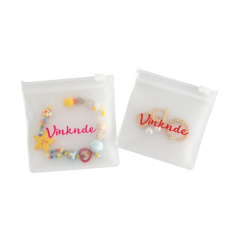 Puede incluir: Dos bolsas de pl&aacute;stico transparente con cierre de cremallera con la palabra "Vinknde" impresa en rosa en cada bolsa. Una bolsa contiene un brazalete de cuentas de colores con un dije de estrella y la otra bolsa contiene un anillo de oro y un pendiente de perla.