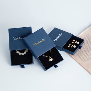 Op de afbeelding: Drie blauwe doosjes met gouden opschrift "Vinknde" en een zwart fluwelen interieur. De doosjes bevatten een parelketting, een parel armband en parel oorbellen.