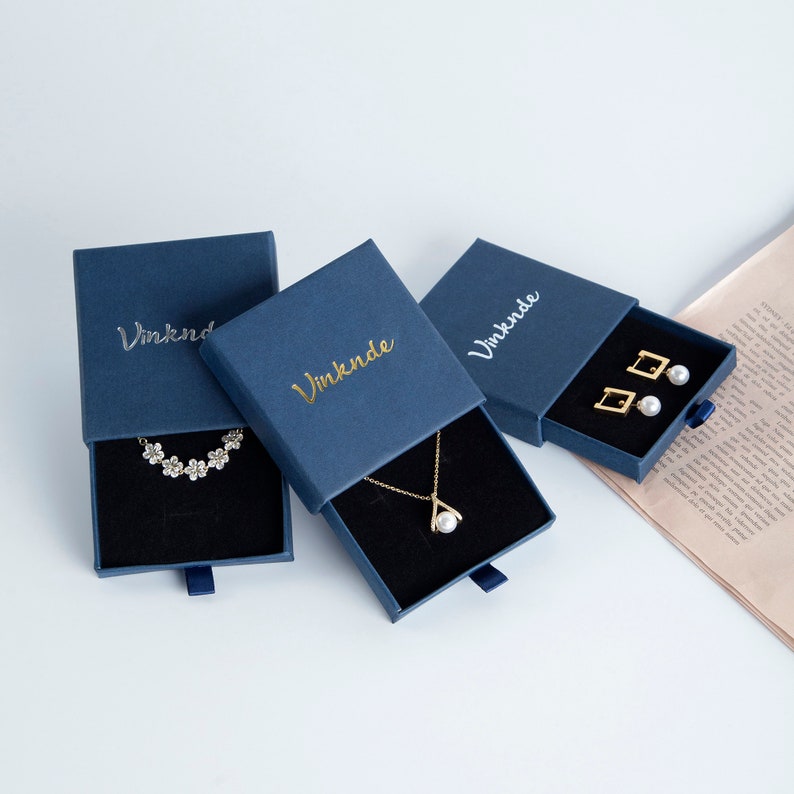 Op de afbeelding: Drie blauwe doosjes met gouden opschrift "Vinknde" en een zwart fluwelen interieur. De doosjes bevatten een parelketting, een parel armband en parel oorbellen.