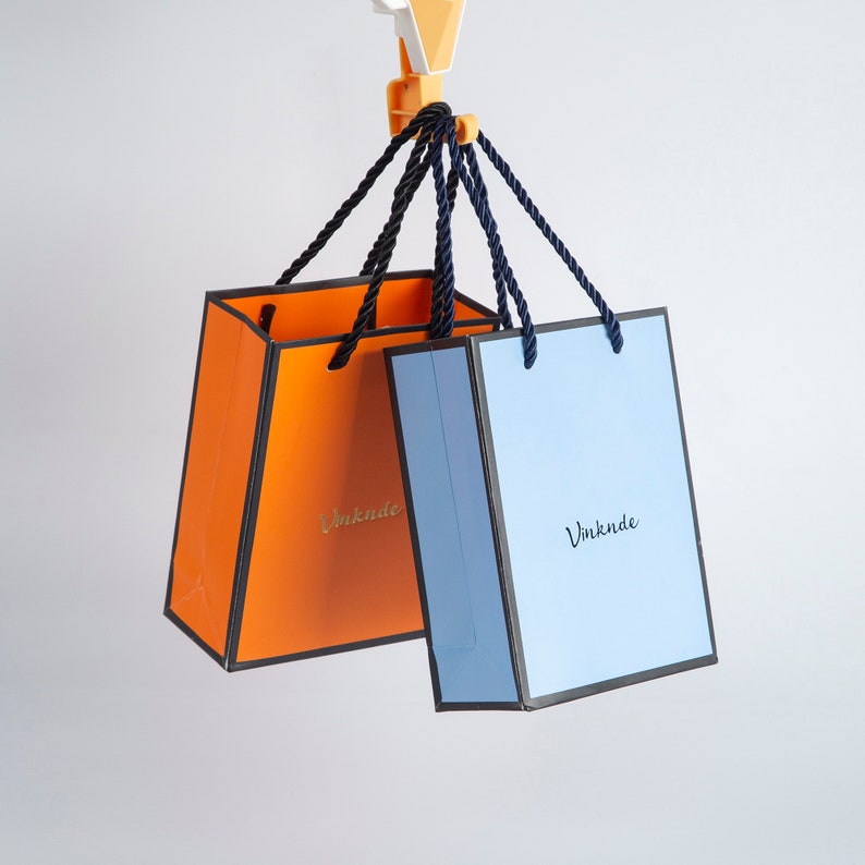 May include: Two shopping bags hanging from a hook. The bag on the left is orange with black trim and the text "Vinknde" in gold lettering. The bag on the right is blue with black trim and the text "Vinknde" in gold lettering.