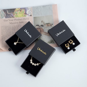 Op de afbeelding: Drie zwarte geschenkdozen met schuifladen, elk met een gouden inscriptie "Vmknde" en een ander sieraad. De eerste doos bevat een parelketting, de tweede een ketting met bloemen en de derde een paar gouden oorbellen met parel accenten.