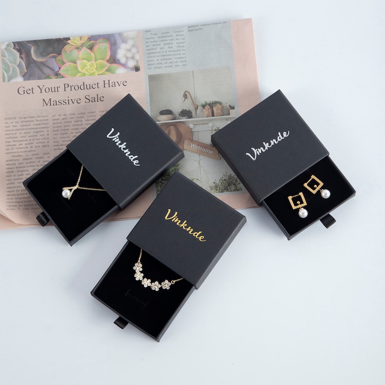 Op de afbeelding: Drie zwarte geschenkdozen met schuifladen, elk met een gouden inscriptie "Vmknde" en een ander sieraad. De eerste doos bevat een parelketting, de tweede een ketting met bloemen en de derde een paar gouden oorbellen met parel accenten.
