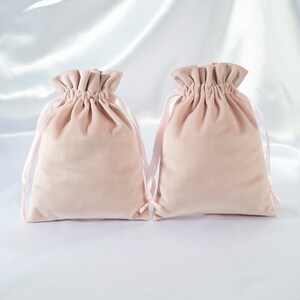Pode incluir: Duas bolsas de cord&atilde;o de veludo rosa claro com la&ccedil;os de fita de cetim rosa. As bolsas est&atilde;o sobre um fundo de cetim branco.