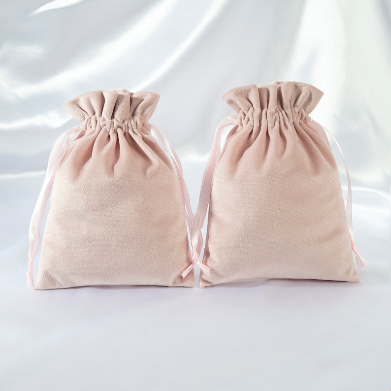 Pode incluir: Duas bolsas de cord&atilde;o de veludo rosa claro com la&ccedil;os de fita de cetim rosa. As bolsas est&atilde;o sobre um fundo de cetim branco.