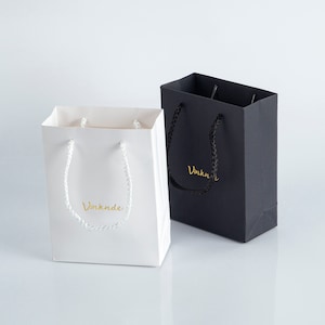 May include: Two small gift bags, one white and one black, with white rope handles. Both bags have the word "Vinknde" printed in gold on the front.
