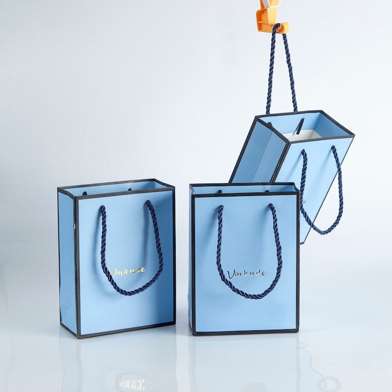 May include: Three light blue gift bags with black trim and navy blue rope handles. The bags have the text "VinKode" printed in gold on the front.