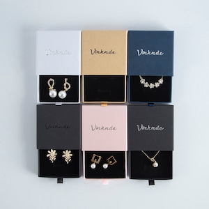 Op de afbeelding: Zes kleine geschenkdozen met een zwart fluwelen interieur. Elke doos heeft een andere kleur aan de buitenkant: wit, bruin, blauw, zwart, roze en zwart. De dozen bevatten sieraden: parel oorbellen, een bloemenketting, bloemen oorbellen, vierkante oorbellen en een parelketting. De dozen zijn gelabeld met "Vinknde".