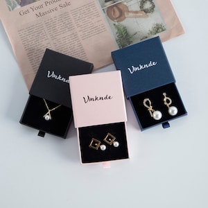 Op de afbeelding: Drie kleine geschenkdozen met lades, elk in een andere kleur: zwart, roze en blauw. De dozen hebben het woord "Vinknde" op de bovenkant gedrukt. Elke doos bevat een sieraad: een ketting met een parel hanger, een paar oorbellen met parel studs en een paar oorbellen met parel hangertjes.