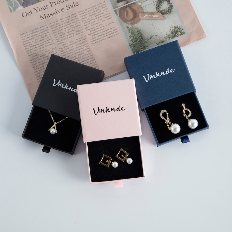 Op de afbeelding: Drie kleine geschenkdozen met lades, elk in een andere kleur: zwart, roze en blauw. De dozen hebben het woord "Vinknde" op de bovenkant gedrukt. Elke doos bevat een sieraad: een ketting met een parel hanger, een paar oorbellen met parel studs en een paar oorbellen met parel hangertjes.