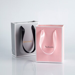 May include: Two gift bags with ribbon handles. The bag on the left is gray with black ribbon handles and the text "Vinknde" in gold lettering. The bag on the right is pink with pink ribbon handles and the text "Vinknde" in gold lettering.