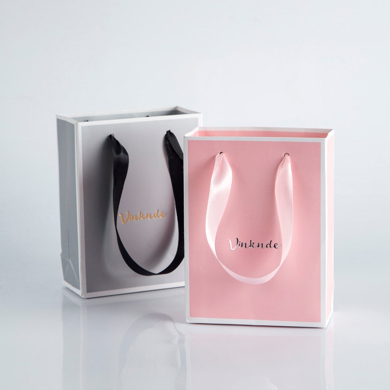 May include: Two gift bags with ribbon handles. The bag on the left is gray with black ribbon handles and the text "Vinknde" in gold lettering. The bag on the right is pink with pink ribbon handles and the text "Vinknde" in gold lettering.