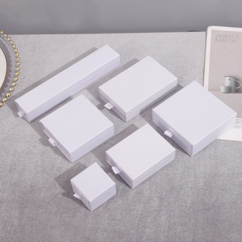 May include: Six white cardboard boxes with sliding lids. The boxes are of various sizes and are arranged on a gray surface.