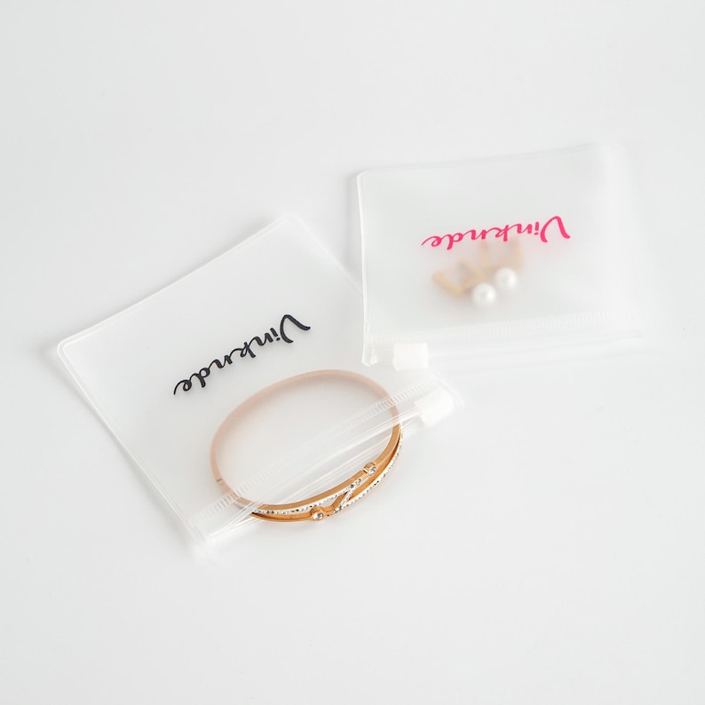 Puede incluir: Dos bolsas de pl&aacute;stico transparentes con cierre de cremallera. Una bolsa contiene una pulsera de oro con un dise&ntilde;o delicado. Ambas bolsas tienen la palabra "remarkable" impresa, una en negro y la otra en rosa.