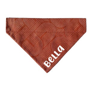 Op de afbeelding: Een roestkleurige bandana voor huisdieren met een wit, retro-achtig patroon van gebogen lijnen. Het woord "Bella" is in witte, gestileerde letters in de rechterbenedenhoek gedrukt. De bandana heeft een driehoekige vorm.