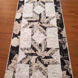 May include: A black and white table runner with a floral pattern. The runner features a geometric design with a repeating star pattern. The runner is approximately 14 inches wide and 36 inches long.