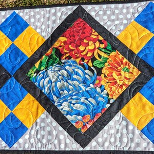 May include: A quilted table runner with a black diamond-shaped center featuring a floral print in shades of red, yellow, blue, and green. The diamond is surrounded by a gray and white polka dot fabric with black stitching. The runner has a blue, yellow, and gray border.