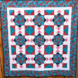 May include: A colorful quilt with a repeating geometric pattern. The quilt features squares and triangles in teal, red, black, and white. The border is teal with a floral pattern. The quilt is displayed against a wooden background.