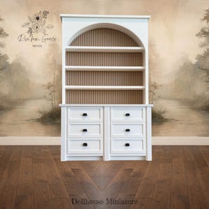 May include: A white dollhouse miniature hutch with a curved top and vertical wooden slats in the center. It has three shelves and six drawers with black knobs. The words "Rise from Grace MINIS" are above the hutch.