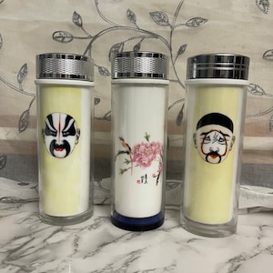 May include: Three glass tea tumblers with silver lids. Two tumblers feature yellow backgrounds with painted faces, while the center tumbler has a white background with a floral and bird design. The tumblers are cylindrical.