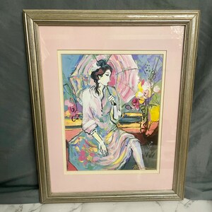 Könnte beinhalten: Gerahmtes Kunstwerk mit einer sitzenden Figur, die einen rosa Sonnenschirm hält. Das Kunstwerk hat eine Pastellfarbpalette mit rosa, blauen und gelben Farbtönen. Der Rahmen ist silberfarben und das Kunstwerk ist rosa mattiert.