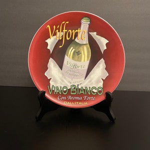 Può includere: Un piatto decorativo con bordo rosso e immagine centrale di una bottiglia di vino bianco. Il piatto presenta la scritta "Vilforte" in oro e "Vino Bianco" in verde, con testo aggiuntivo "Con Aroma Forte" e "Dall'Italia".