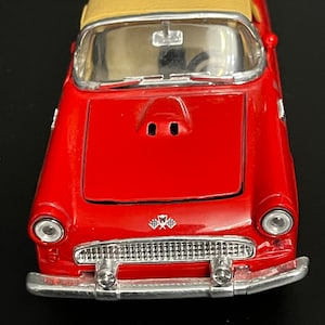 Puede incluir: Un coche de juguete rojo con techo descapotable beige. El coche tiene detalles cromados en el parachoques delantero y los faros. Un emblema de bandera a cuadros es visible en la parte delantera. Es un modelo clásico.