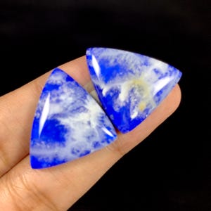 May include: Two triangular, polished gemstones with a vibrant blue hue and white marbling. The gemstones are held against a dark background, showcasing their glossy surface and unique patterns. These stones are likely used for jewelry or decorative purposes.