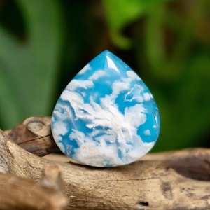 May include: A teardrop-shaped cabochon gemstone featuring a vibrant sky-blue base color with intricate, swirling white inclusions that resemble clouds. The polished surface is smooth and reflective, showcasing a distinct, organic pattern.