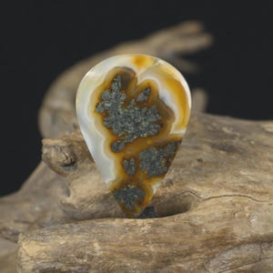 May include: A teardrop-shaped gemstone with a distinctive pattern. The stone displays a mix of white, yellow, and brown hues, with dark grey inclusions. The gemstone is set against a natural wood background.